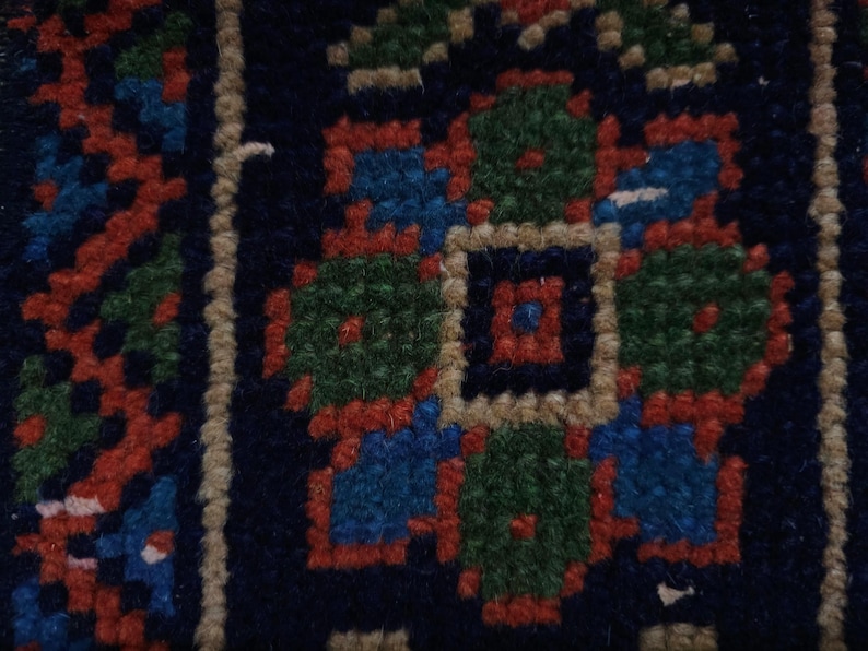 Op de afbeelding: Close-up van een deel van een donkerblauw tapijt met een herhalend bloemmotief. Het patroon heeft groene, rode en blauwe bloemmotieven met een beige rand. Het tapijt heeft een getextureerd uiterlijk.
