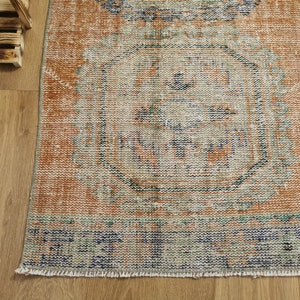 May include: A vintage-style rug with a faded orange background and a blue and green geometric pattern. The rug has a fringed edge.