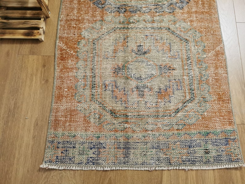 May include: A vintage-style rug with a faded orange background and a blue and green geometric pattern. The rug has a fringed edge.