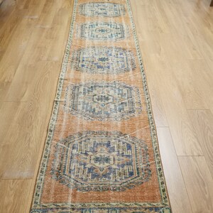May include: A long, rectangular rug with a faded orange background and a blue and white geometric pattern. The rug has a fringed edge.
