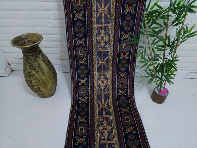 Op de afbeelding: Een lang, patroonrijk tapijt met een donkerblauwe achtergrond en ingewikkelde geometrische ontwerpen in tinten goud, rood en groen. Een decoratieve vaas en een plant in pot staan ook in de afbeelding.