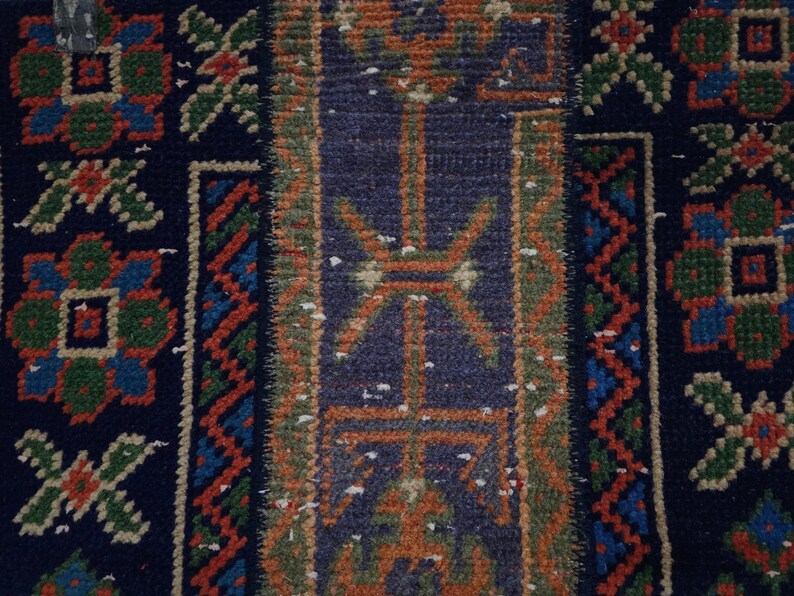 Op de afbeelding: Close-up van een deel van een patroonkleed. Het kleed heeft een donkerblauwe achtergrond met een centraal paneel in een lichter blauw en oranje geometrisch ontwerp. De randen hebben bloemen- en zigzagpatronen.