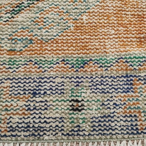 May include: A close-up of a woven rug with a geometric pattern in shades of brown, blue, green, and white. The rug has a fringe border.
