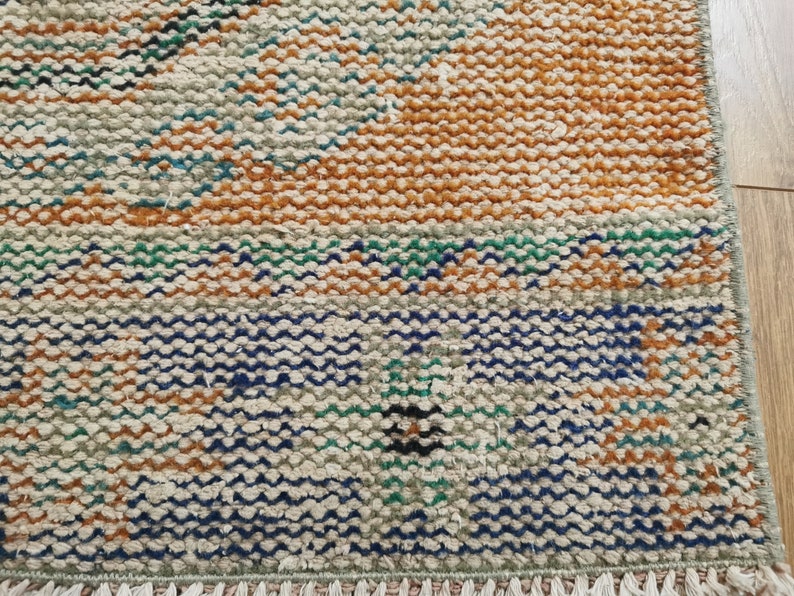 May include: A close-up of a woven rug with a geometric pattern in shades of brown, blue, green, and white. The rug has a fringe border.