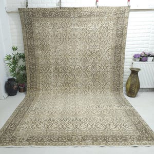 May include: A large rectangular area rug featuring a repeating intricate floral and vine pattern in shades of beige and muted brown. The rug has a wide decorative border with a similar botanical motif, creating a symmetrical design throughout.