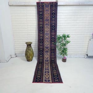 Op de afbeelding: Een lang, rechthoekig tapijt met een centraal patroon van geometrische vormen in tinten blauw, goud en rood. Het tapijt wordt verticaal tentoongesteld tegen een witte bakstenen muur. Een decoratieve vaas en een kleine plant staan op de voorgrond.