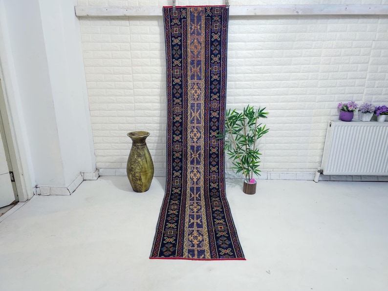 Op de afbeelding: Een lang, rechthoekig tapijt met een centraal patroon van geometrische vormen in tinten blauw, goud en rood. Het tapijt wordt verticaal tentoongesteld tegen een witte bakstenen muur. Een decoratieve vaas en een kleine plant staan op de voorgrond.