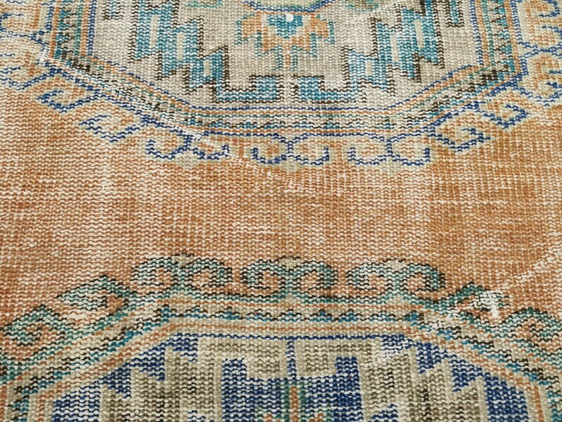 May include: A close-up of a vintage rug with a faded orange background and intricate blue and brown patterns. The rug has a worn and distressed look, adding to its character.