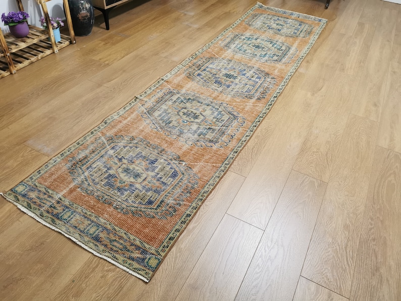 May include: A long, rectangular rug with a faded orange background and intricate blue and green patterns. The rug has a fringed edge and is laid out on a wooden floor.