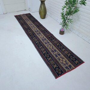 Op de afbeelding: Een lange, smalle loper met een gedetailleerd geometrisch patroon. Het tapijt heeft een donkerblauwe achtergrond met een centraal ontwerp in beige, bruin en paars. De randen zijn rood afgewerkt.