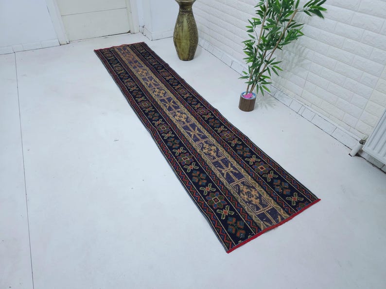 Op de afbeelding: Een lange, smalle loper met een gedetailleerd geometrisch patroon. Het tapijt heeft een donkerblauwe achtergrond met een centraal ontwerp in beige, bruin en paars. De randen zijn rood afgewerkt.