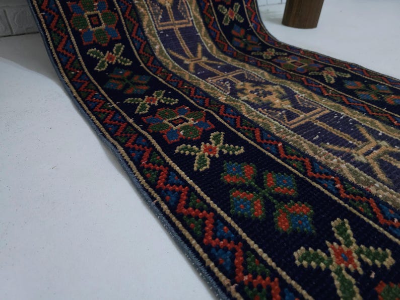 Op de afbeelding: Detailopname van een marineblauw loperkleed met gedetailleerde patronen. Het kleed heeft een herhalend ontwerp van bloemen en geometrische vormen in rood, groen en goud tegen een donkerblauwe achtergrond. De randen zijn afgewerkt met een blauwe rand.
