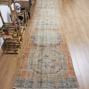 May include: A vintage runner rug with a faded, distressed look. The rug has a light orange background with a geometric pattern in shades of blue, beige, and orange. The rug is approximately 2.5 feet wide and 8 feet long.