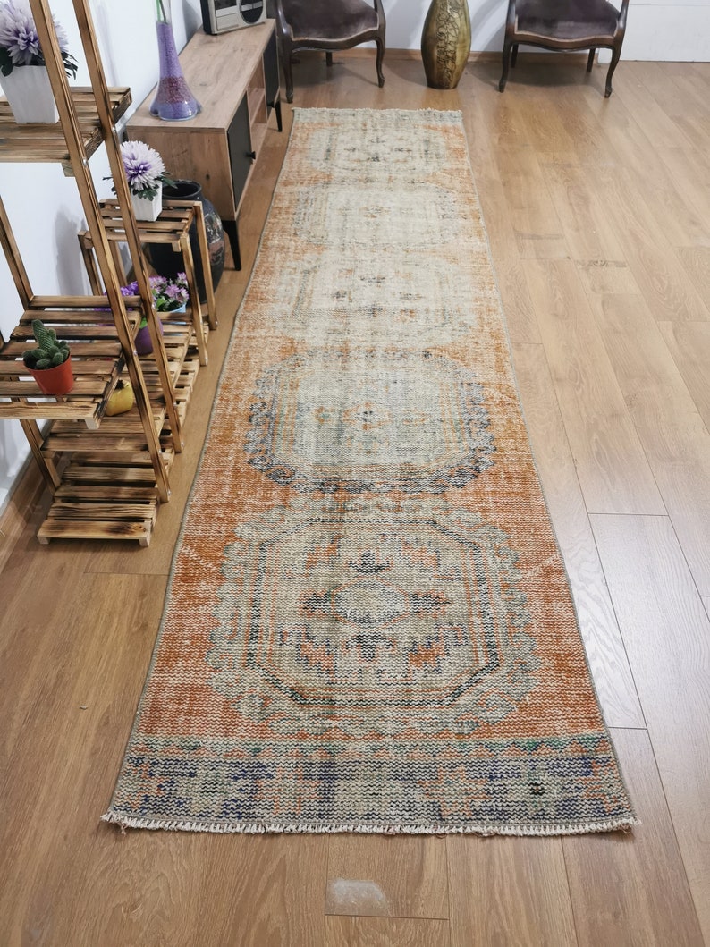 May include: A vintage runner rug with a faded, distressed look. The rug has a light orange background with a geometric pattern in shades of blue, beige, and orange. The rug is approximately 2.5 feet wide and 8 feet long.