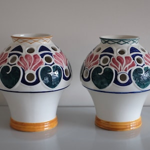 May include: Two white ceramic vases with a floral design. The vases have a yellow rim and are decorated with blue, green, and pink flowers. The vases have holes in the sides for flowers.