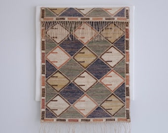 May include: A woven rug with a geometric pattern of diamonds in shades of brown, beige, green, and blue. The rug has a fringe border.