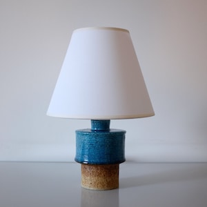 以下が含まれることがあります： 白いランプシェードが付いた青と茶色のセラミック製のテーブルランプ。ランプはテクスチャのあるベースと円筒形のシェードを持っています。