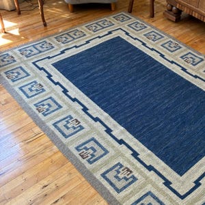 May include: A rectangular area rug featuring a solid deep blue center panel surrounded by a wide border. The border displays a repeating geometric pattern in shades of light beige, blue, and brown, creating a structured frame around the rug.