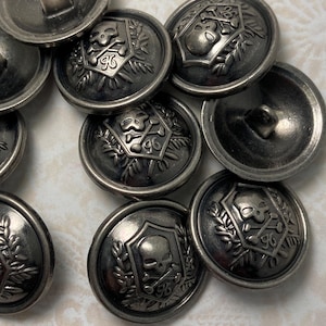 May include: A collection of antique silver-colored metal buttons. Each button features a skull and crossbones design within a shield, surrounded by a laurel wreath and the number 96. The buttons have a slightly tarnished, vintage appearance.