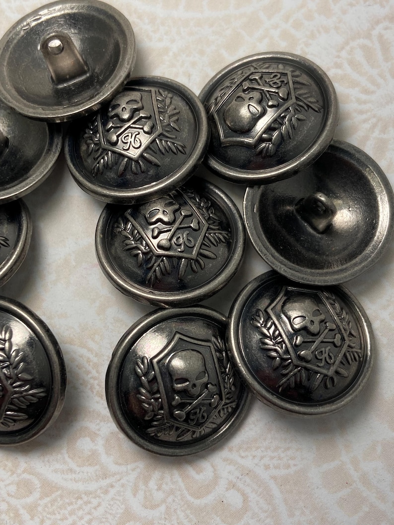 May include: A collection of antique silver-colored metal buttons. Each button features a skull and crossbones design within a shield, surrounded by a laurel wreath and the number 96. The buttons have a slightly tarnished, vintage appearance.