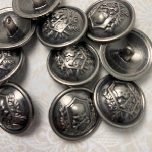 May include: A collection of antique silver-toned metal buttons. Each button features a detailed embossed design, including shields, laurel wreaths, and other decorative elements. The buttons have a slightly tarnished appearance, suggesting age.