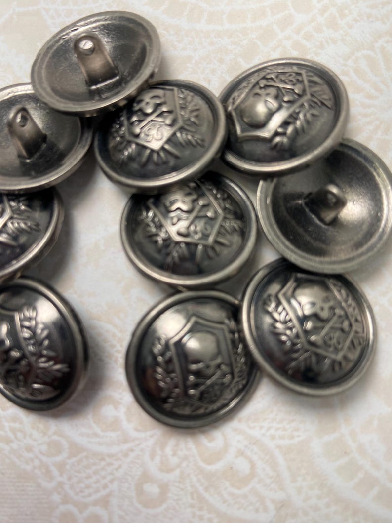 May include: A collection of antique silver-toned metal buttons. Each button features a detailed embossed design, including shields, laurel wreaths, and other decorative elements. The buttons have a slightly tarnished appearance, suggesting age.