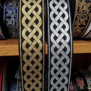 May include: Two rolls of decorative ribbon with a Celtic knot design. One ribbon is black with gold metallic accents, and the other is black with silver metallic accents. The ribbons are displayed on a wooden shelf.
