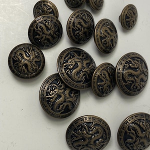 May include: A collection of antique bronze-colored buttons with a dragon design. The buttons vary in size and feature a detailed relief of a dragon within a circular frame. The buttons have a dark, aged appearance.