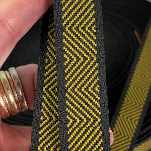 Könnte beinhalten: Schwarz-goldenes Band mit Fischgrätmuster. Das Band hat einen schwarzen Hintergrund mit einem sich wiederholenden goldenen Zickzack-Design. Das Band ist dicht gewebt und scheint haltbar zu sein. Eine Rolle des Bandes ist im Hintergrund sichtbar.