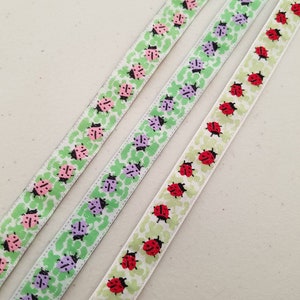 Può includere: Tre nastri decorativi con motivi a coccinella. Un nastro ha coccinelle rosa, un altro ha coccinelle viola e il terzo ha coccinelle rosse. Tutti i nastri hanno un motivo a foglie verdi su sfondo bianco.