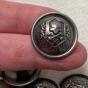 May include: Silver-toned metal button with a skull and crossbones design inside a shield, surrounded by laurel leaves. The number "96" is below the skull. The button has a raised edge and a slightly tarnished appearance.