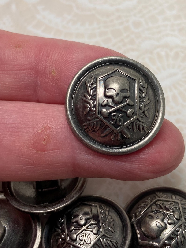 May include: Silver-toned metal button with a skull and crossbones design inside a shield, surrounded by laurel leaves. The number "96" is below the skull. The button has a raised edge and a slightly tarnished appearance.