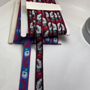 Peut inclure: Ruban décoratif avec des motifs floraux rouges, blancs et verts sur fond noir. Un ruban bleu avec des motifs floraux rouges et blancs est également visible. Les rubans sont empilés dans un contenant en carton blanc.