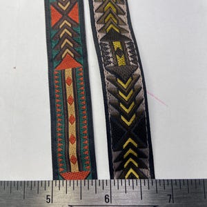 May include: Two decorative ribbons with geometric patterns. One ribbon features a design in orange, green, and brown. The other ribbon has a design in black, yellow, and brown. A ruler is placed below the ribbons.