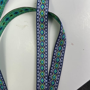 May include: Close-up of decorative ribbon in blue, green, black, and white geometric patterns. The ribbon features a repeating diamond design with a contrasting border. The ribbon is likely used for crafting or embellishment.