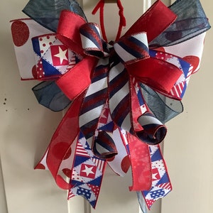 May include: A red, white, and blue bow with a patriotic theme. The bow is made of various ribbons with different patterns, including stars, polka dots, and stripes. The bow is tied with a red ribbon and is hanging from a hook.