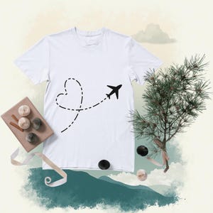 Puede incluir: Camiseta blanca con un diseño de avión negro y una trayectoria en forma de corazón punteada. La camiseta se muestra sobre un fondo neutro con elementos decorativos, incluyendo un pequeño árbol y regalos envueltos. Un diseño sencillo con temática de viajes.