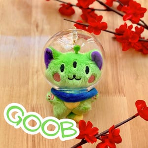 May include: A plush green alien toy with three eyes, purple ears, and a blue collar is encased in a clear bubble. The word "GOOB" is written in green and white on the bottom left. Red flowers and branches are in the background.