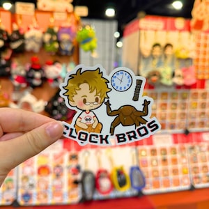 Puede incluir: Una pegatina con un personaje de dibujos animados, un reloj y el texto "CLOCK BROS". La pegatina tiene un borde blanco y se muestra frente a un puesto de mercado.