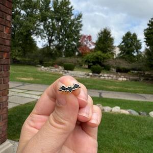May include: A small, black bat-shaped enamel pin with a gold-colored border. The pin is held between fingers against a backdrop of green grass, trees, and a cloudy sky. The pin is a Halloween-themed accessory.