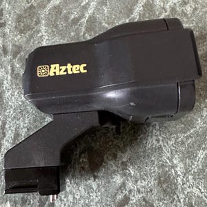 Puede incluir: Un flash de cámara Aztec negro. El flash tiene un cuerpo rectangular con un cabezal giratorio y un soporte de montaje. El logotipo de Aztec está en letras doradas. La unidad está diseñada para usarse con una cámara.
