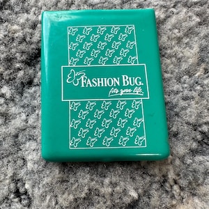 Puede incluir: Un objeto rectangular de color verde azulado con el texto "Fashion Bug. fits your life" en blanco. El objeto tiene un acabado brillante y presenta un patrón de ilustraciones de mariposas blancas.
