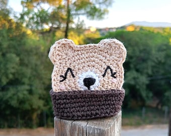 Może przedstawiać: Szydełkowana podstawka pod kubek w kształcie misia z jasnobrązową twarzą, wyszywanymi czarnymi oczami i białym pyszczkiem z czarnym nosem. Dolna część wykonana jest z ciemnobrązowej włóczki, tworząc kontrastującą obwódkę wokół głowy misia.