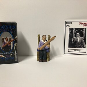 May include: A small figurine of John Stanley, a comic book artist, sitting on a throne with a large pencil in his hand. The figurine is made of clay and is painted in realistic colors. The figurine is packaged in a box with the words "Phoenix Comics & Toys" and "John Stanley Mini Figurine". A tribute card with a black and white photo of John Stanley is also included.