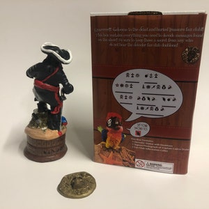 May include: A pirate figurine with a black coat, red sash, and white hat stands on a wooden base with a compass design. The figurine is holding a sword. The background is a red and white box with a pirate parrot and the text "Gyarrrrr!!! Welcome to the dead and buried treasure fan club! This box contains everything you need to decode messages found on the show! Be sure to keep these a secret from any who do not bear the decoder fan club doubloon!" and a code chart.