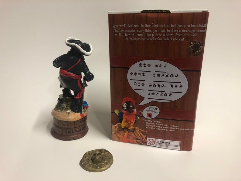 May include: A pirate figurine with a black coat, red sash, and white hat stands on a wooden base with a compass design. The figurine is holding a sword. The background is a red and white box with a pirate parrot and the text "Gyarrrrr!!! Welcome to the dead and buried treasure fan club! This box contains everything you need to decode messages found on the show! Be sure to keep these a secret from any who do not bear the decoder fan club doubloon!" and a code chart.