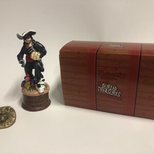 May include: A detailed figurine of a pirate holding a bag of gold coins, standing on a barrel with a parrot and skull. The figurine is part of a Phoenix Comics Buried Treasures collection. The figurine is packaged in a wooden box with the Phoenix Comics logo and the words "Buried Treasures".