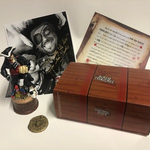 May include: A wooden treasure chest box with a pirate figurine, a gold coin, and a black and white photo of a pirate with a signature. The box is labelled "The Captain's Buried Treasures" and the photo is signed "Ahoy Matey!"