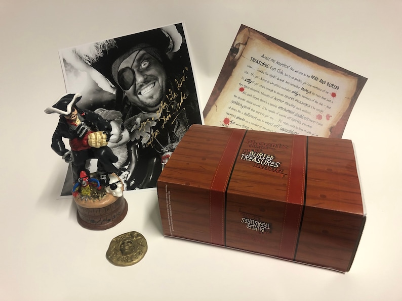 May include: A wooden treasure chest box with a pirate figurine, a gold coin, and a black and white photo of a pirate with a signature. The box is labelled "The Captain's Buried Treasures" and the photo is signed "Ahoy Matey!"