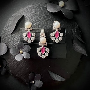 May include: A set of silver earrings and a pendant featuring a pink gemstone surrounded by clear stones. The earrings and pendant are on a black display stand.